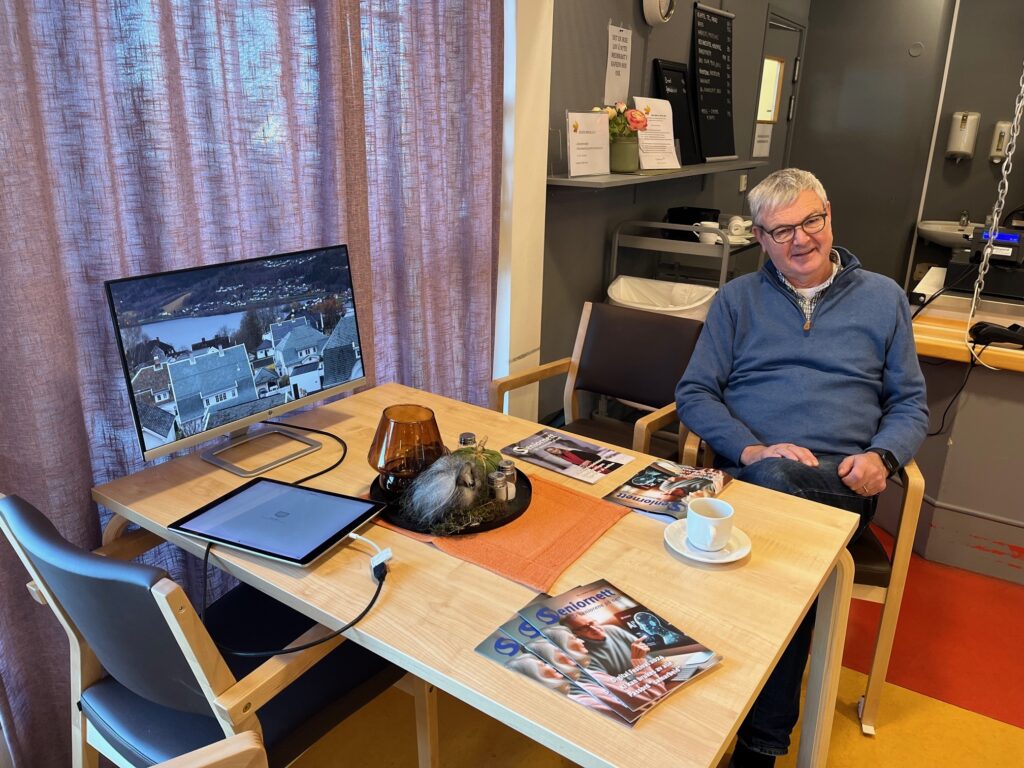 på et bord står en skjerm og viser digitale bilder, mann sitter ved siden klar til å veilede