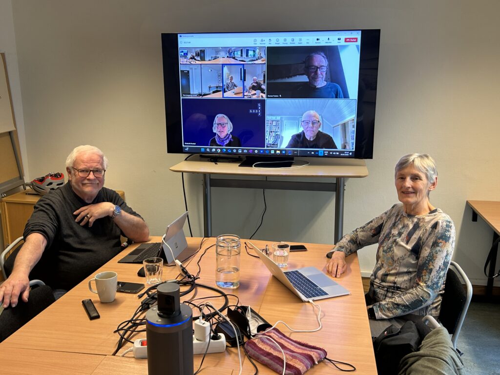En mann og en kvinne sitter ved et bord hvor tre mennesker vises på en skjerm.
